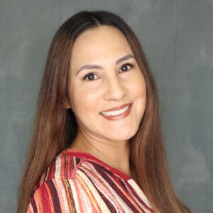 Tabitha is smiling while looking over her right shoulder. She has long brown straight hair. She is wearing a red, white, and brown striped long sleeve shirt.
