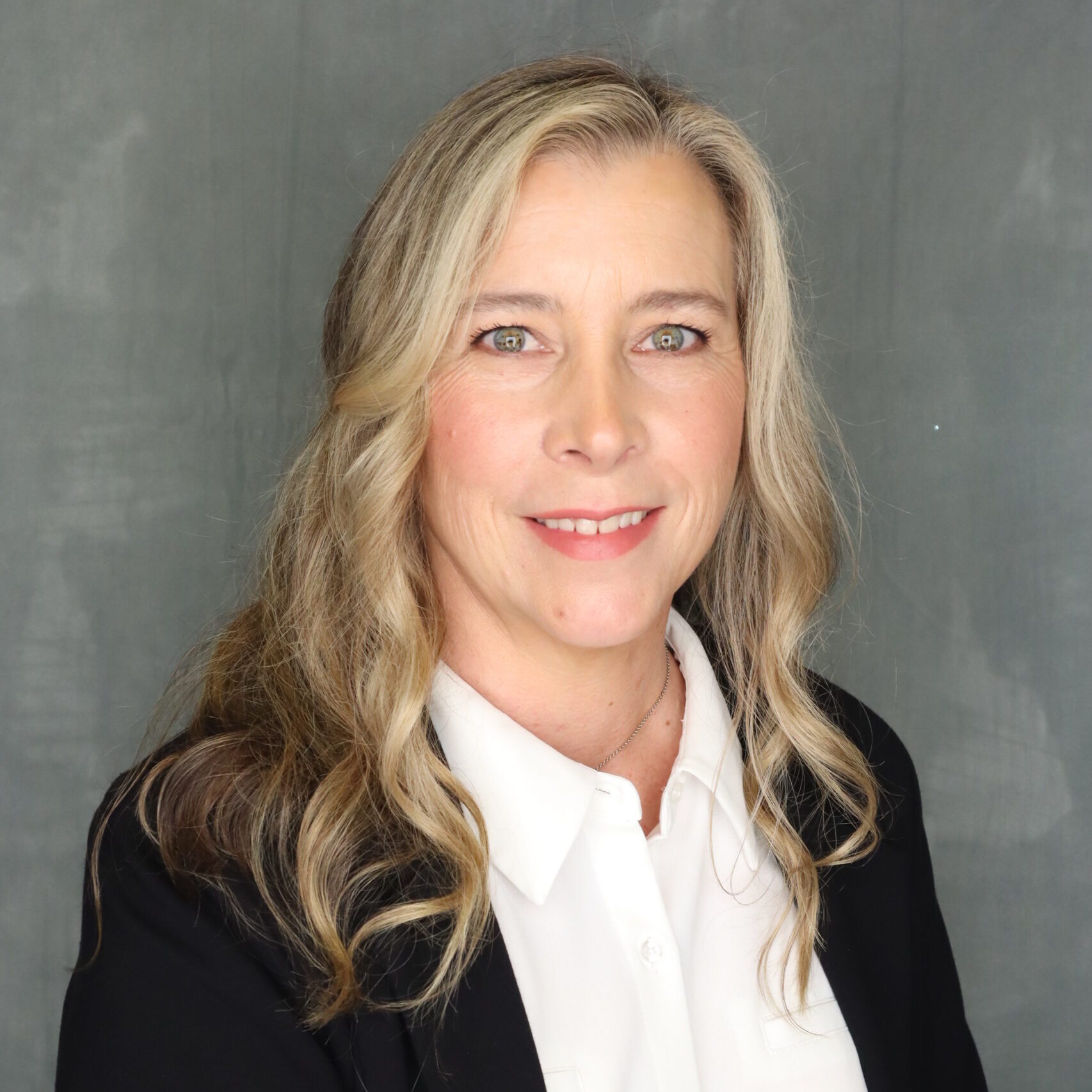 Kit is smiling. She has long blonde curled hair. She is wearing a black blazer with a white button up shirt underneath.
