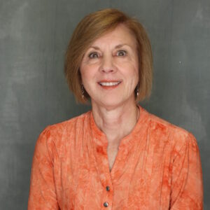 Lynne is smiling. She has short blonde hair. She is wearing an orange long sleeve shirt. She is wearing earrings with the Power T on them.
