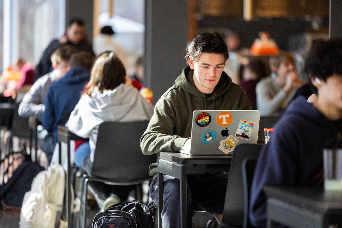 A student works on a laptop while studying in a busy campus dining hall