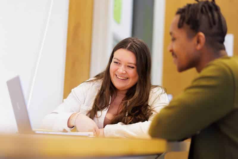 Two students smiling and talking across a table with a laptop open between them