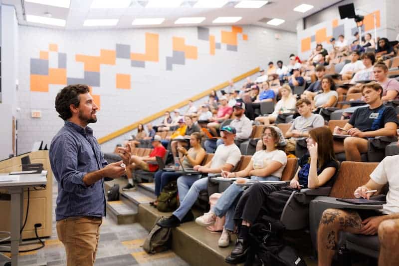 Instructor speaking to a full class of students seated in a tiered lecture hall