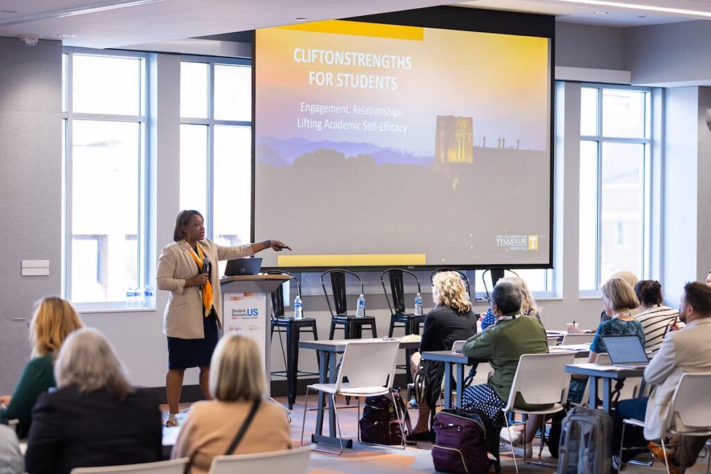 Presenter pointing to a projected slide while attendees watch from tables at a conference session