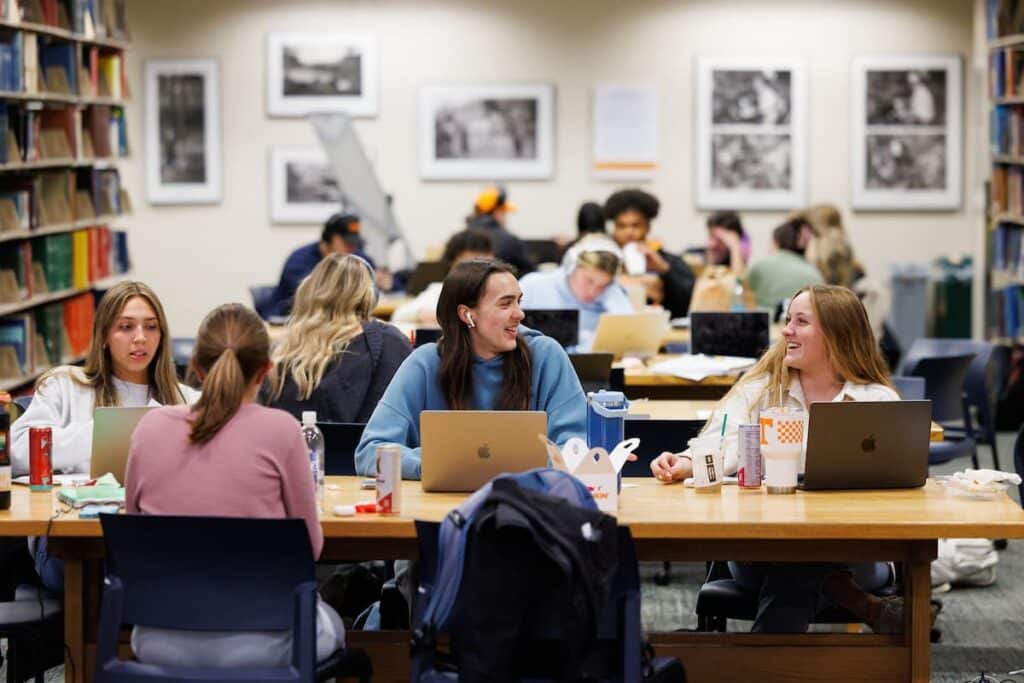 Students studying and talking over laptops at long tables in a busy library