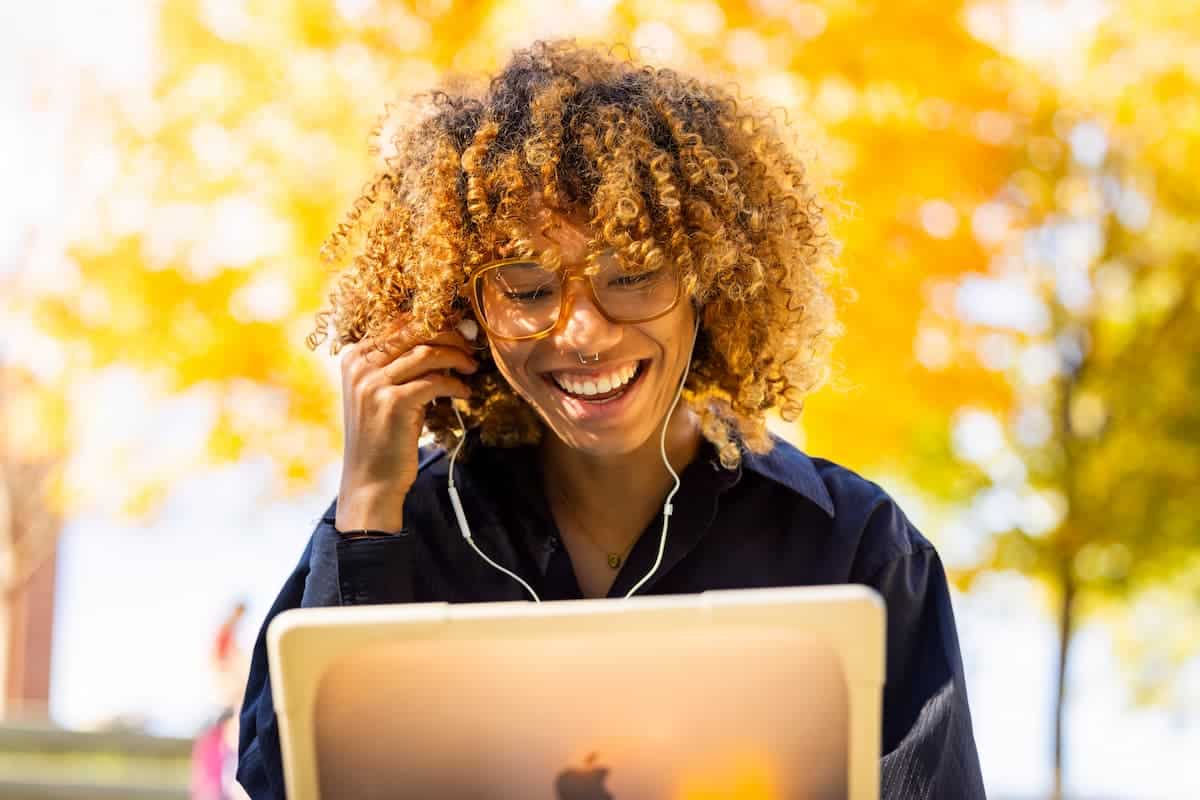 Student smiles at a laptop screen while wearing earbuds outdoors under fall foliage