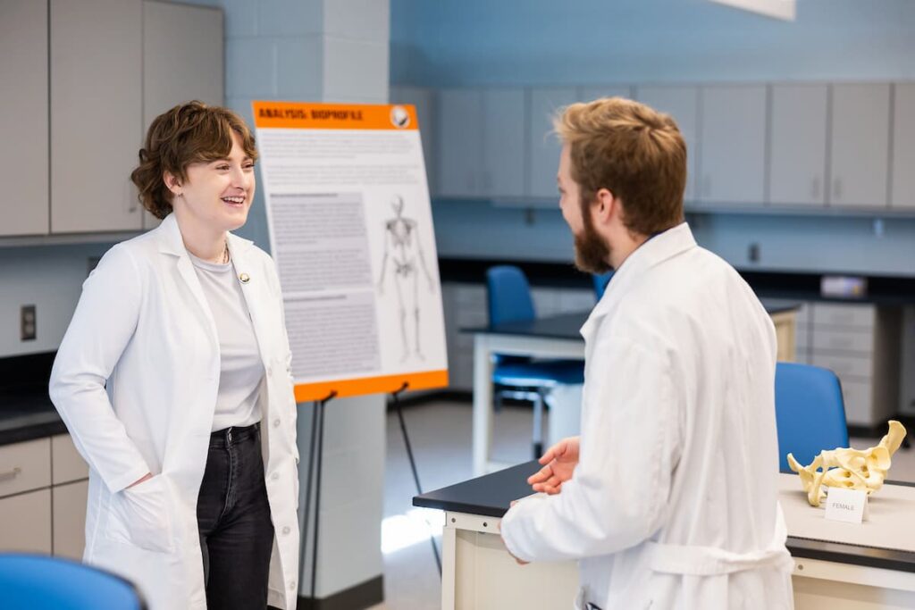 Two researchers in white lab coats talk beside a research poster and skeletal specimens in a lab