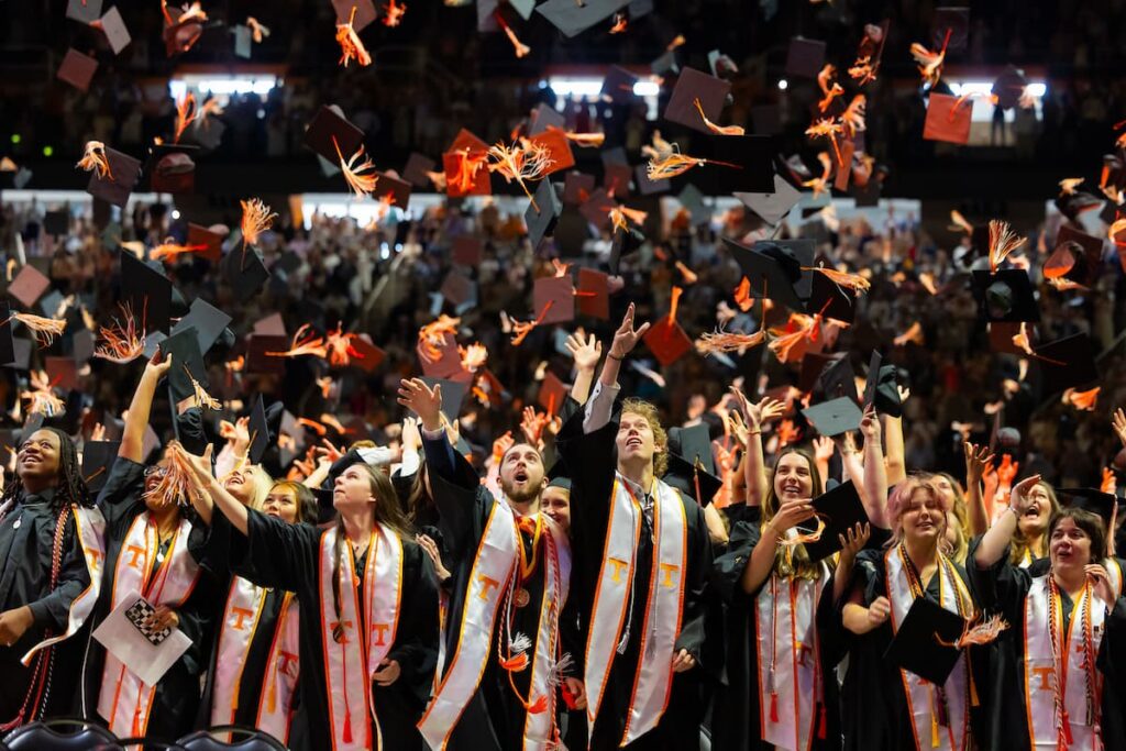 Graduates in Power T stoles throw their caps skyward inside a packed commencement arena