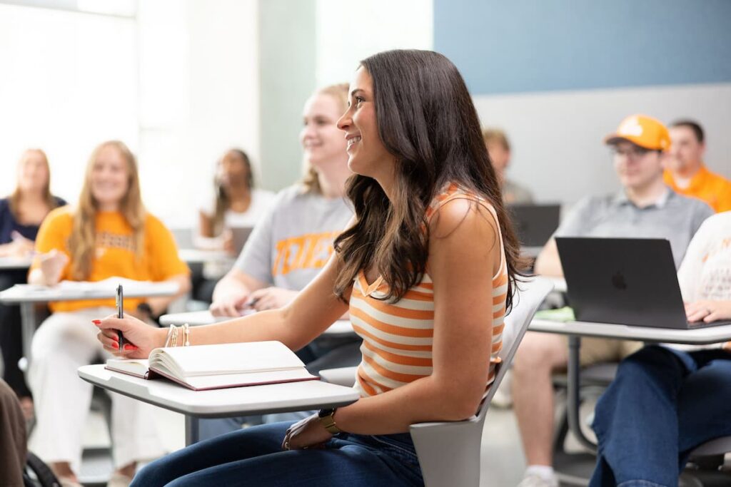 Student smiles while taking notes at a desk during a class, with classmates seated behind