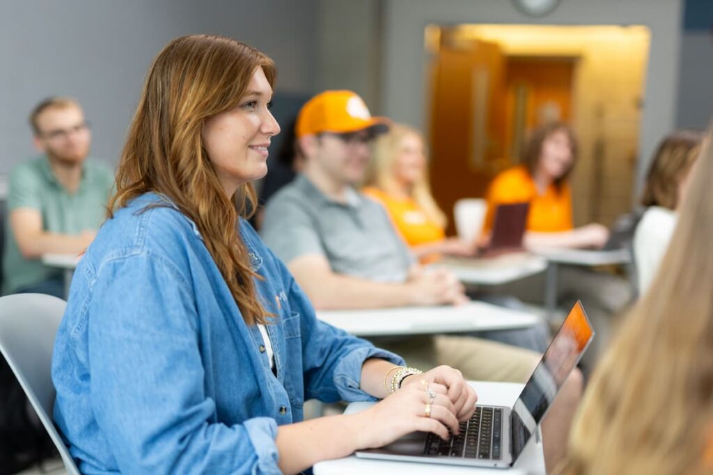 Student typing on a laptop during a class session, with classmates seated at desks behind her