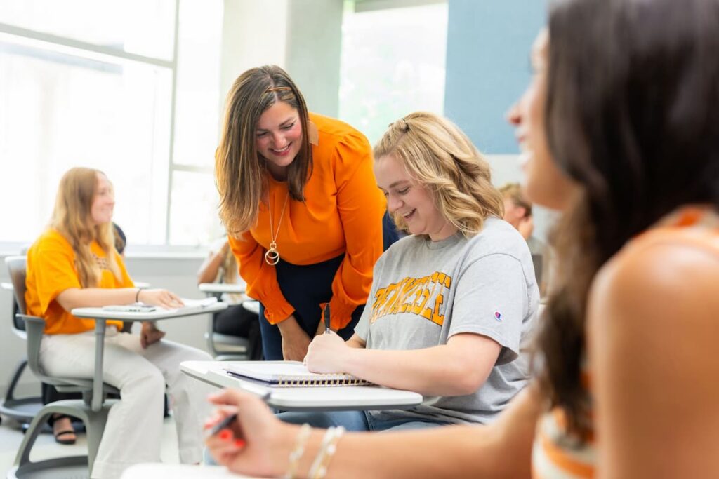 Instructor leaning over a classroom desk to help a student taking notes