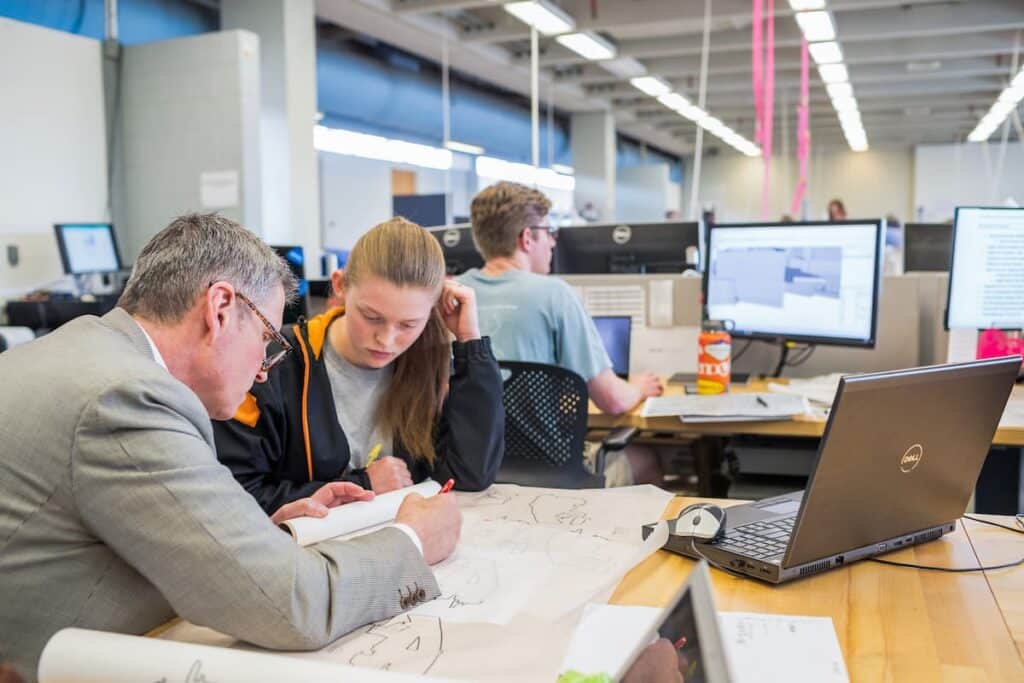 Faculty member and student reviewing hand-drawn plans at a computer workstation in a design studio