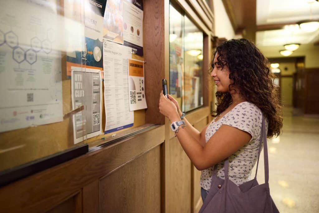 Student photographing a flyer on a hallway bulletin board with her phone