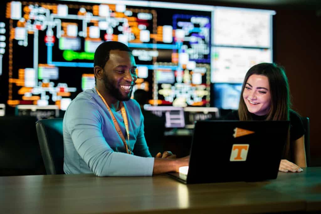 Two students smiling at a laptop in front of a large wall of data visualization screens