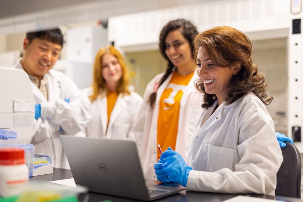 Researcher in a lab coat working at a laptop while three lab members look on in a research lab