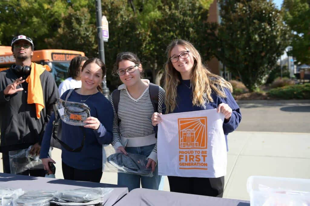 Three students at an outdoor table holding up a Proud to be First Generation T-shirt