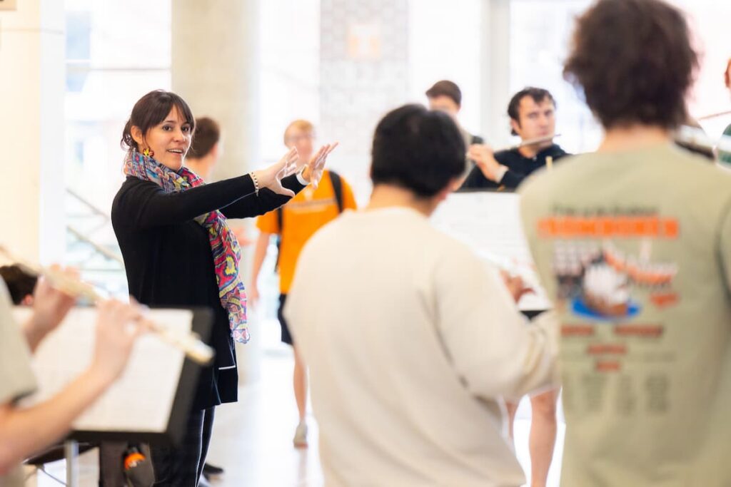 Instructor directing a group of student musicians rehearsing with flutes in a sunlit campus atrium