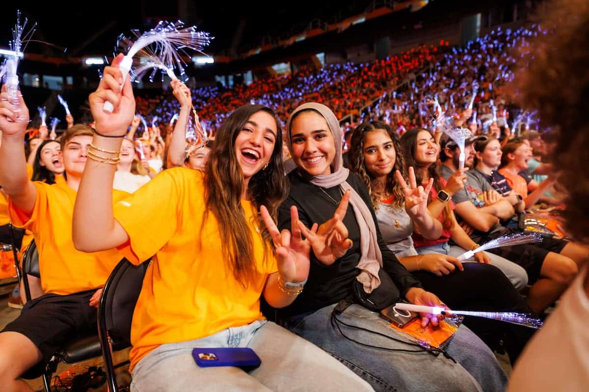 Students in packed arena seating cheering with light-up wands and raised hand signs