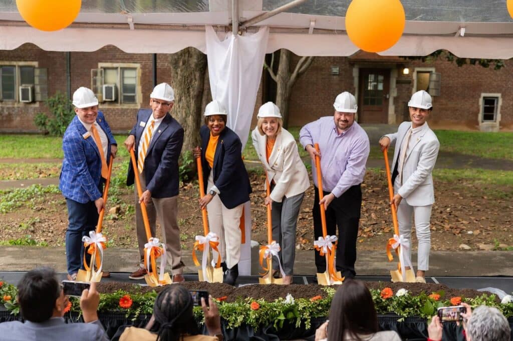 Six people in hard hats turning dirt with ceremonial orange shovels at a groundbreaking ceremony